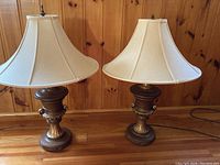 Pair of decorative table lamps with fabric shades and bronze-toned ornate bases placed on wooden floor against a wood-paneled wall.