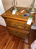 Wood nightstand with two drawers, top drawer with carved floral design and brass ring pull, bottom drawer with brass handle, set on hardwood floor with two metal lamps on top.