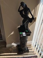 Full view of black glossy female figurine statue mounted on fluted column pedestal base near window with sunlight casting shadows.