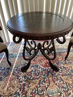 Front angle view of the round wooden table showing patterned embossed center, curved legs, and nailhead trim edge.