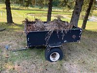 Side view of metal framed utility trailer with wood sides filled with yard debris, showing large off-road tires and black paint with wear.