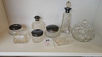 Full view of six antique cut glass items: perfume bottles, candy bowl, trinket bowl, dresser jars arranged on a shelf against a neutral striped wallpaper background, including a small paper label '89'.