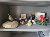 Shelf with multiple bird figurines including bronze bird on stone, several smaller ceramic birds, and bird with red scarf on pine branch, plus two framed miniature watercolors.