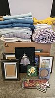 Stack of assorted textile linens and blankets in various colors alongside multiple framed artworks and picture frames arranged together