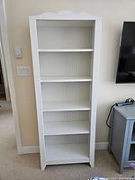 Front view of tall white wooden book shelf with five shelves and decorative top edge, showing overall condition.