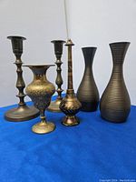 Front view of two brass candlestick holders, two ribbed brass vases, ornate brass vase, and brass rose water sprinkler on blue surface.
