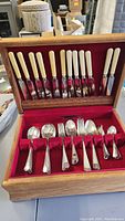 Full view of flatware set neatly arranged in wooden box with knives displayed in the lid section and spoons and forks in the base section lined in red velvet.