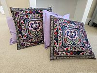 Four toss cushions including two with intricate colorful embroidered floral and bird patterns on black fabric and two solid light purple cushions.