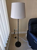 Full view of the floor lamp showing the black turned spindle base, beige fabric shade, power cord and foot switch beside a black recliner chair.