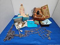 Photo of various vintage items displayed on blue cloth including hat pins, pocket watch, box with chess pieces, lipstick holder mirror, sequined necklace, and other small vintage collectibles.