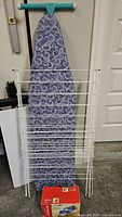 Front view of the turquoise Seymour ironing board with patterned fabric cover, white metal laundry drying rack in front, and boxed Sunbeam iron on the floor.