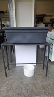 Metal plant stand with rectangular black plastic planting pot placed on top and white ceramic pot on the ground beneath