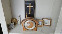 Wide photo of shelf showing vintage Smiths mantel clock centered with two small framed watercolours on right, framed cross artwork in back, and other decor including floral hanging and plaque on left.
