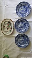 Four plates laid out on white cloth: three blue and white Spode Italian plates with architectural scenes and one multicolor Ashworth platter with floral and bird motifs