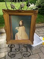 Full view of the framed George Baxter print displayed on a decorative metal stand outdoors, showing the entire print and frame with visible corner and edge wear.