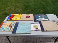 Seven 78 RPM record albums and records displayed on a white folding table outdoors on grass and pavement. Various colorful album covers visible including two Roy Rogers albums.