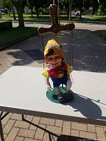Front view of the Pinocchio puppet with its colorful clothing, sitting on the green base stand outdoors on a table.