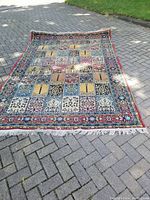 Top view of the colorful Asian carpet laid on a paved surface, showing geometric and floral square panels with fringe edges, colors include red, blue, beige, black, and gold.
