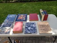 Eight books displayed on a table outdoors, featuring titles on occult art, spiritualism, history, and classic literature including 'Grimoires & Alchemies' and 'The Family Mark Twain'.