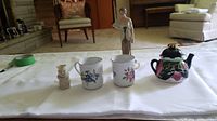 Overview of all items in the lot displayed on a white surface, including figurine, mugs, toby jug, and cat teapot