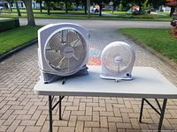 Photo showing both Airworks fans on a table outdoors, one large box fan and one smaller desk fan.