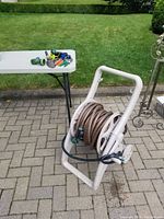 Wide shot showing white plastic hose reel with a beige garden hose wound around it on paved outdoor surface, with green grass and garden bushes in background.