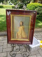 Photo showing the full view of the double framed George Baxter print The Bridesmaid on a black metal art easel outdoors with greenery in background.