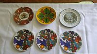 Six plates displayed on white cloth: Three Villeroy & Boch Acapulco plates with colorful birds and flowers; Poole dish with green and brown floral design; Prattware 19th century rural scene plate; Royal Worcestershire Golden Jubilee coat of arms plate.
