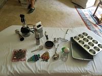Kitchen lot items laid out on white tablecloth showing blender, baking tins, tins, utensils and kitchen magnets.