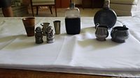 Full view of all items including wooden cup, metal cream and sugar set, two flasks, measures, and figurines on white cloth.