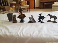 Photo showing the full collection of six metal figures displayed on a white cloth including cup, bust, unicorn scene, knight, horse, and seated figure.