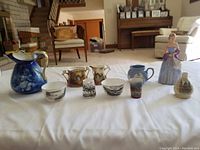 Full view of the nine miscellaneous pieces including cream and sugar sets, jugs, cups and figurines on a white cloth