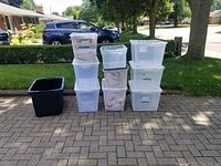 Front view of all 9 storage containers stacked on a brick driveway including translucent plastic containers with lids and two large black bins without lids