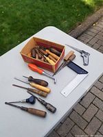 Photo showing miscellaneous hand tools arranged on a white table outdoors including chisels, screwdrivers, a metal try square, and other tools.