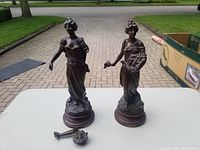 Two spelter statues on wooden pedestals shown, one with detached arm and floral bouquet lying beside it.