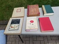 Six vintage books displayed on a white table outdoors, including two large Courier & Ives volumes, The Life of Henry VIII by Antonia Fraser, a red hardcover, a green and cream hardcover, and a marbled red spine hardcover book.