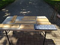 Seven wooden signs displayed on a plastic folding table outdoors on pavement with grass in background. Various branded wine-themed signs and one humorous fishing-themed sign visible.