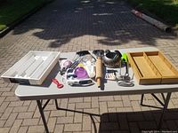 Wide shot of all kitchen items laid out on a table outside including the rolling pin, mashing tool, steamer, measuring spoons, peeler, small utensils, and two drawer organizers.