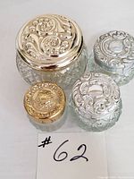Photo of four collectible vanity jars with intricate silver and gold tone lids showing detailed embossed floral and scroll designs in various sizes.