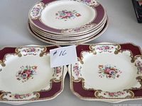Eight pieces of Myott Staffordshire Royal Crown china plates including five round dinner plates stacked and two square plates in front, decorated with floral bouquet center and burgundy gold borders.