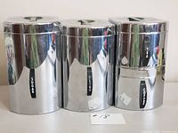 Front view of four shiny metal canisters labeled Sugar, Flour, Tea, and Coffee with black bakelite handles on lids, showing minor surface wear and reflections.
