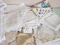 Photo showing folded white and cream linens including embroidered and lace tablecloths, napkins, and doilies in assorted sizes and styles.
