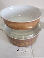 Two vintage Pyrex casseroles stacked, one with clear glass lid, showing the brown Americana pattern and white interior.