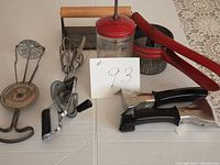 Overview of all vintage kitchen tools including hand-crank chopper, egg beaters, bacon press, red-handled food mill, and Corning casserole handles, laying on white surface