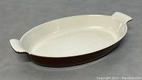 Photo showing the oval cast iron casserole pan with white enamel interior and brown exterior on gray surface, displaying overall shape and color.