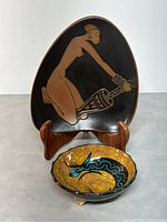Two Kalahari studio pieces, an oval plate on stand with a nude figure design, and a round bowl with antelope stylized artwork, on light surface background.