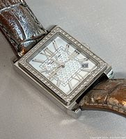 Close-up of Thomas Sabo square faced wristwatch showing crystal bezel, white patterned dial with Roman numerals and date window, silver-tone hands, and brown textured leather strap.