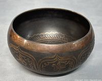 Side angled photo showing the brass singing bowl with detailed Tibetan symbol etching around its outer surface and slight patina wear.