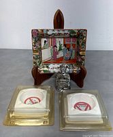 Front view of the Chinese Rose Medallion ashtray displayed on a wooden stand with two white porcelain ashtrays in packaging and a small clear figurine in front