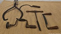 Five antique hand tools including iron ice tongs, brace drill crank, draw kutter, hand sickle, and drill tool with wooden handles on textured mat.